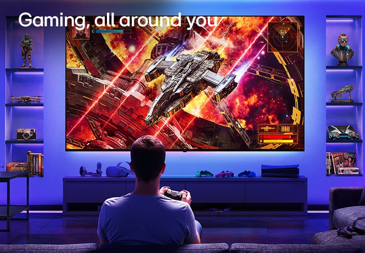 LG Ultra Big TV is wall-mounted in a neon-lit gaming room, where a large display shows a vivid space battle scene as a player sits facing the screen, illustrating an immersive and engaging gaming experience.