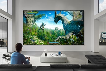 LG TV for gamers appears mounted on a wall in a black-and-white-toned living room, showing a fantasy dinosaur game scene on a large display while a person sits on a sofa holding a controller.