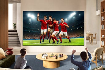 LG TV for sports fans is wall-mounted in a bright living room, showing a soccer celebration with vivid colors and refined picture quality across a wide screen, as a family seated on sofas cheers together.