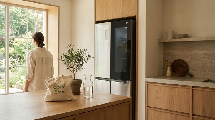 LG refrigerator with sleek glass panel in a natural wood kitchen, beside a large window with greenery and natural light.