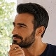 Circulair portret van een man met donker haar en een baard, die zijwaarts kijkt met een nadenkende uitdrukking.