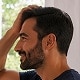 Circulair portret van een man met donker haar en een baard, glimlachend terwijl hij zijn hand door zijn haar haalt.