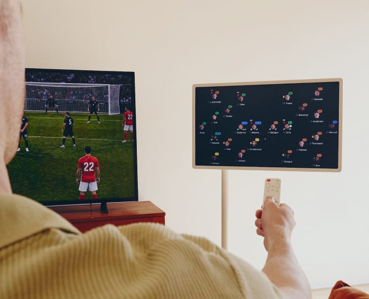 De rug van een man die een afstandsbediening richt op een TV die een voetbalwedstrijd toont en een StanbyME smart screen dat een wedstrijdanalyse weergeeft.