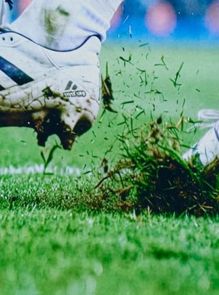Close-up actiebeeld van een voetbalschoen die de grond raakt, waarbij stukken gras en aarde van de grasmat omhoog vliegen.