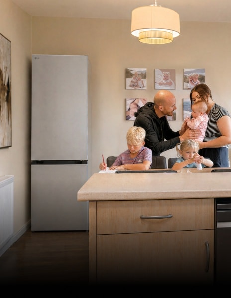 Een gezin verzamelt zich in een keuken bij een LG koelkast – een jongen schrijft aan de eettafel, een jong meisje zit naast hem met een flesje, en ouders staan samen met een baby.