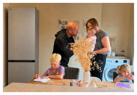 Een gezin verzamelt zich in de keuken bij een roestvrijstalen koelkast – een jongen tekent aan de tafel, een meisje schildert aan het aanrecht, en ouders staan samen met een baby.