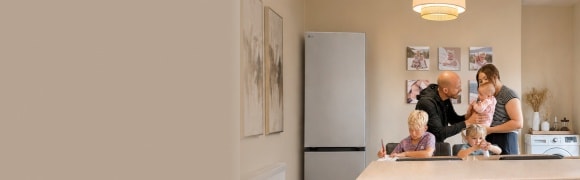 Een gezin met drie kinderen verzamelt zich in een warme, moderne keuken, met de ouders die voor de kinderen zorgen rond een groot keukeneiland. Een koelkast, vaatwasser, oven en keukenkastjes zijn zichtbaar op de achtergrond.