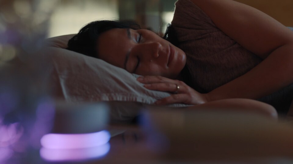 Een vrouw ligt op haar zij in bed te slapen. Voor haar licht het LG ThinQ ON apparaat op. Het gordijn gaat open en een man aait een hond. De vrouw die op haar zij lag wordt wakker.