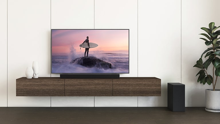 Een LG TV en LG soundbar staan op een bruine plank en de subwoofer staat op de vloer. Het scherm toont een surfer die op de rots staat.