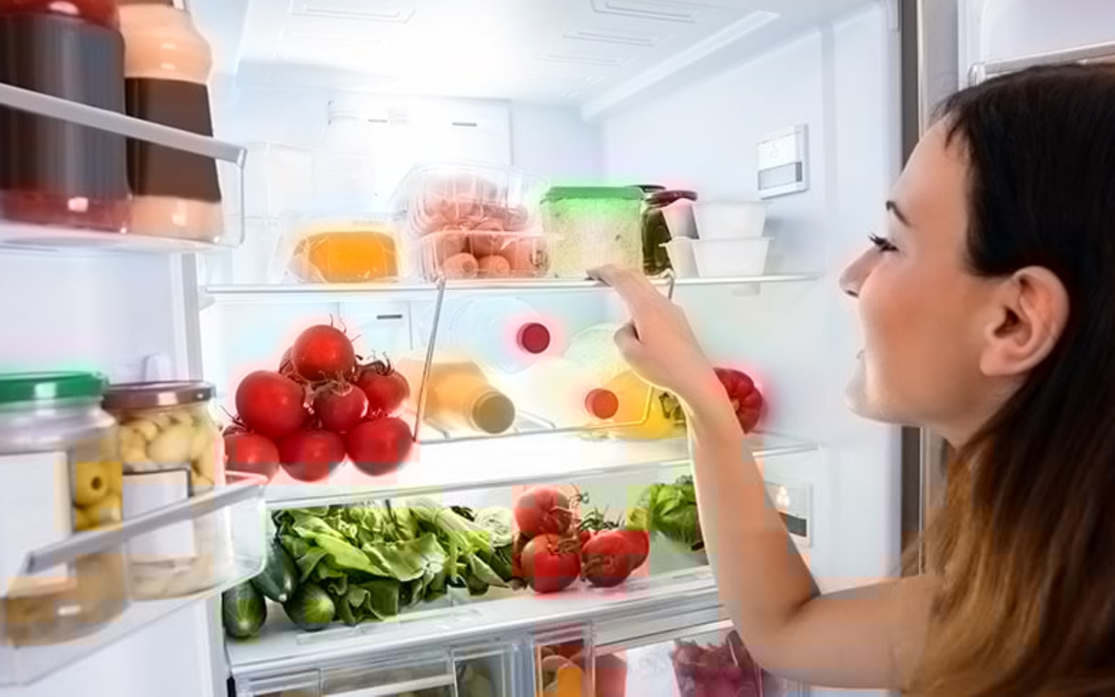 A woman is looking into a fully-stocked fridge filled with tomatoes, vegetables, juices, and more.