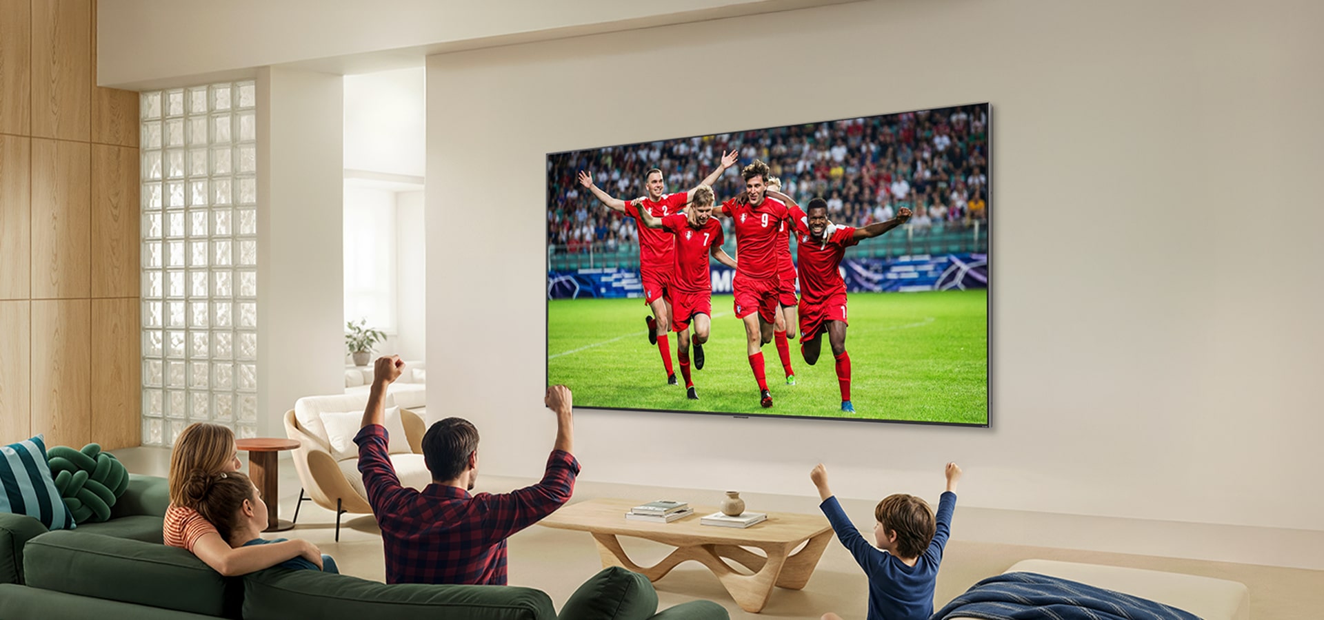 LG QNED AI QNED70 Mini LED Ultra Big TV is wall-mounted in a bright living room, showing an intense soccer moment with vivid colors and refined picture quality across a wide screen, as a family seated on sofas cheers together.