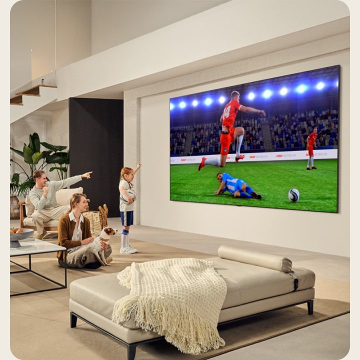 Una familia en la sala de estar. Tienen una LG TV wall mounted con una pantalla ultragrande que es casi tan grande como toda la pared. Están viendo un emocionante partido de fútbol en la gran pantalla inmersiva.  