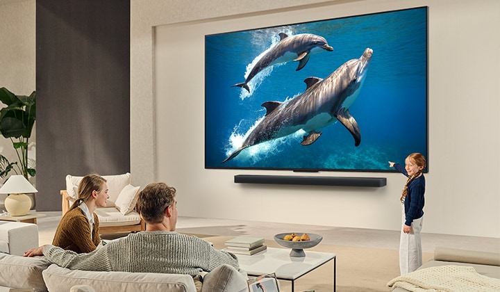 Una familia sentada en el sillón frente a una LG QNED TV montada en la pared; una pequeña niña señala la pantalla que muestra dos delfines. 