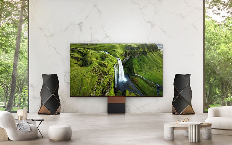 An LG MAGNIT installed on a living room wall, is vividly displaying a large waterfall along with a vast landscape and dynamic sound coming from the Beolab 90 stereo speakers located on both sides of the LG MAGNIT.