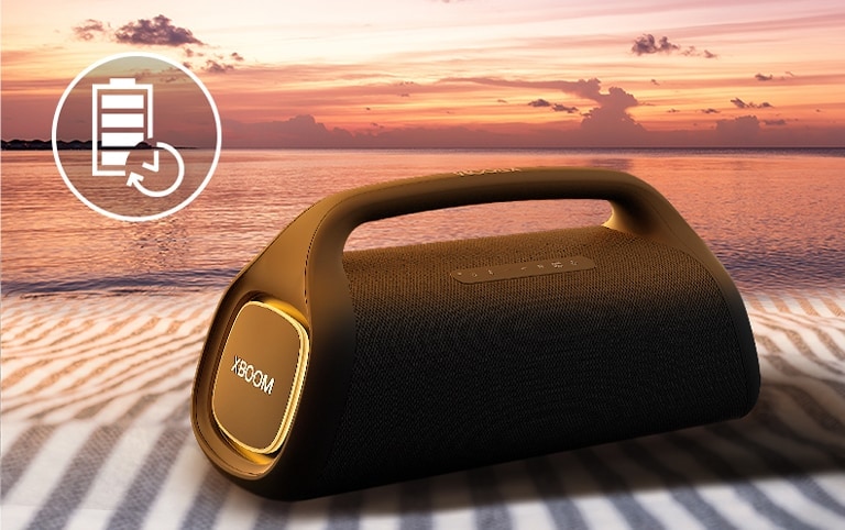 A caixa de som está colocada sobre uma toalha de praia. Na frente dela, vê-se uma praia ao pôr do sol para ilustrar que a caixa de som pode ficar ligada por mais tempo.