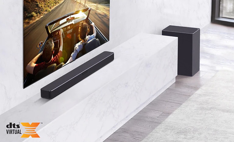 La TV está en la pared, la barra de sonido LG est+a debajo en un estante de mármol blanco con un Subwoofera su derecha. La TV muestra una pareja en un auto.