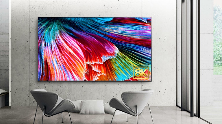 El QNED99 aparece montado contra una pared de hormigón gris con dos sillas grises dispuestas frente al televisor. La pantalla muestra una imagen en primer plano de pétalos de colores brillantes y el logotipo de 8K.