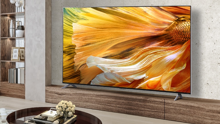 El QNED90 aparece sobre un mueble de madera frente a una pared con la pantalla mostrando una imagen en primer plano de pétalos en tonos amarillos y naranjas.