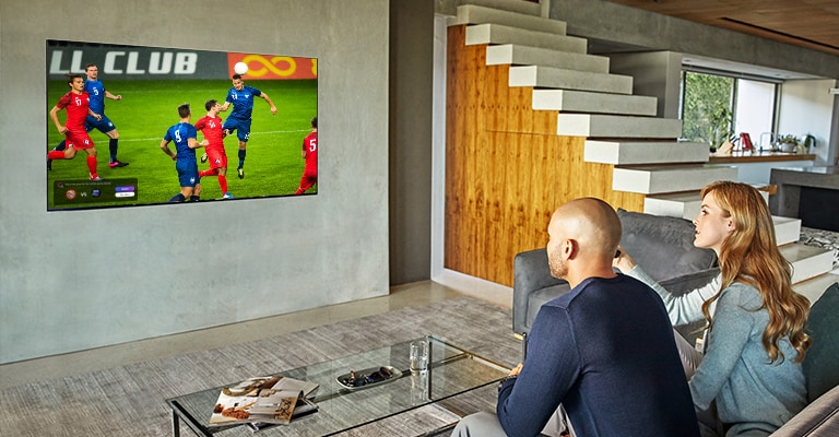 Un hombre y una mujer están sentados detrás de una mesa de café y frente a un televisor de pared que muestra un partido de fútbol.