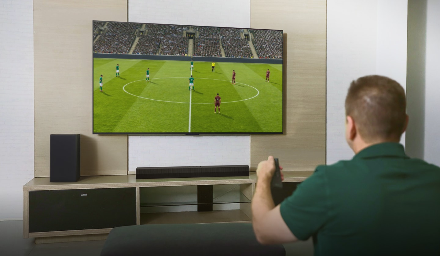 Marcos sostiene un control remoto mientras ve una escena de un partido de fútbol en un televisor grande en la sala.