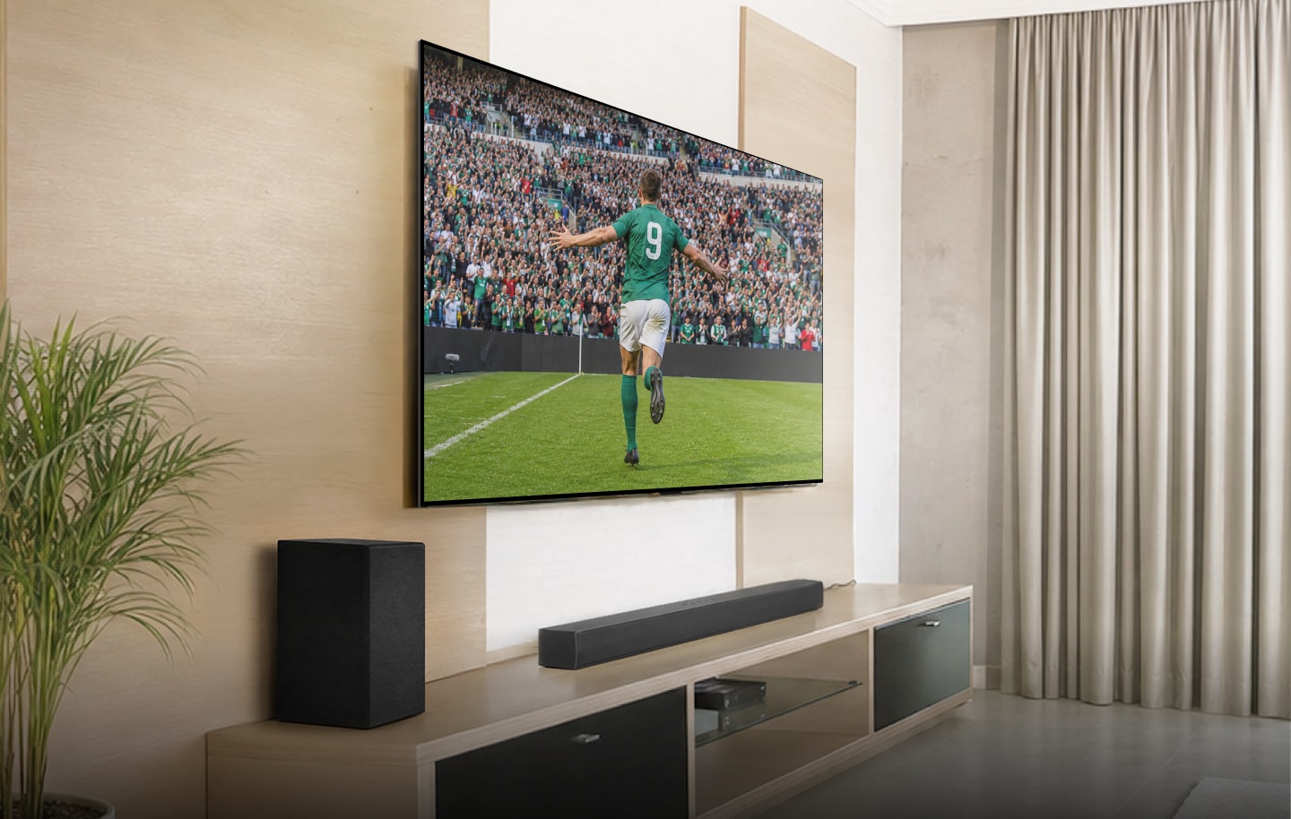 Gran televisor LG montado en la pared con una barra de sonido y un subwoofer, mostrando a un futbolista celebrando un gol.