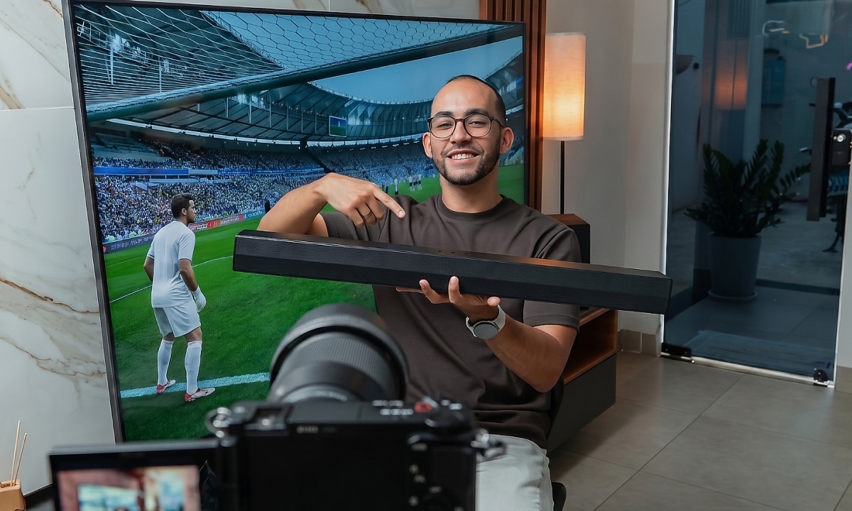 Rapael sonriendo y señalando una barra de sonido que sostiene, con una TV mostrando un partido de fútbol en el fondo