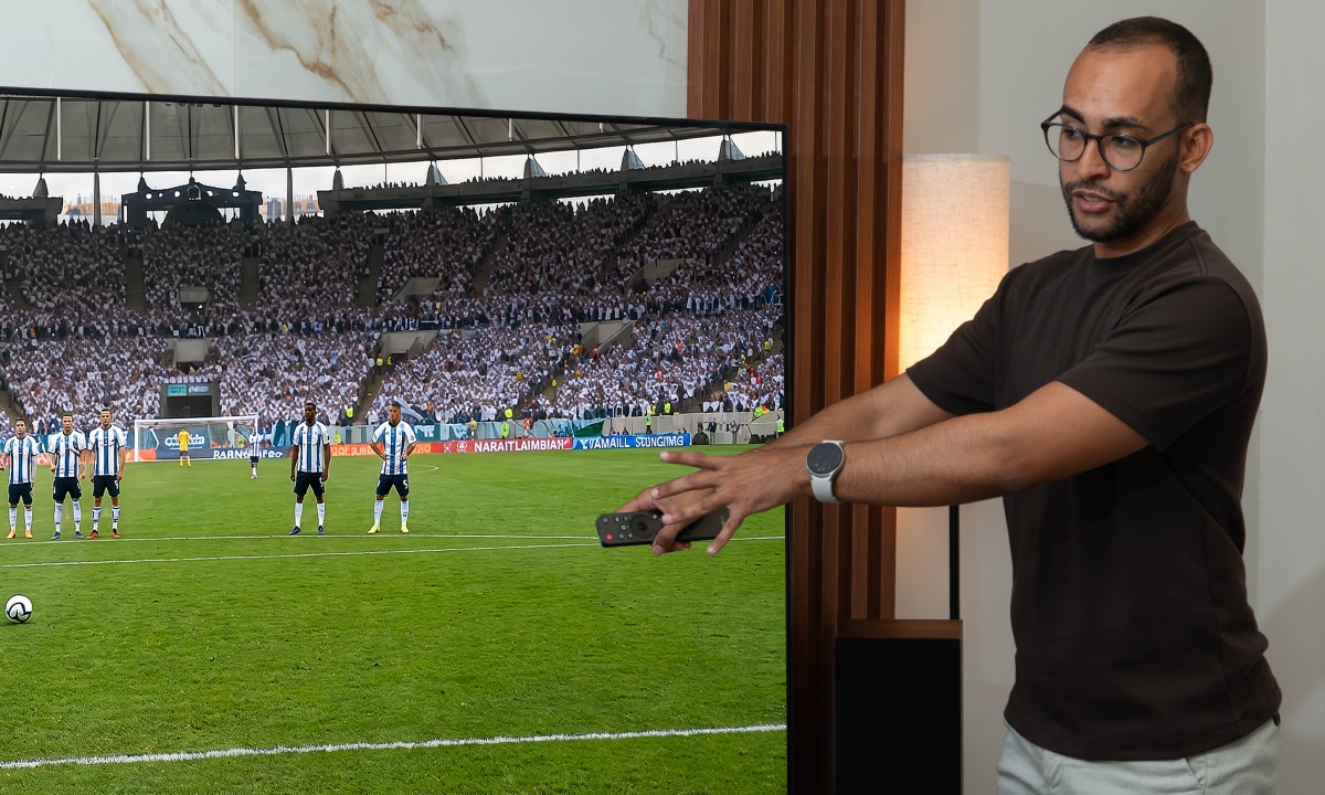 Rapael configurando una barra de sonido usando un control remoto mientras ve un partido de fútbol