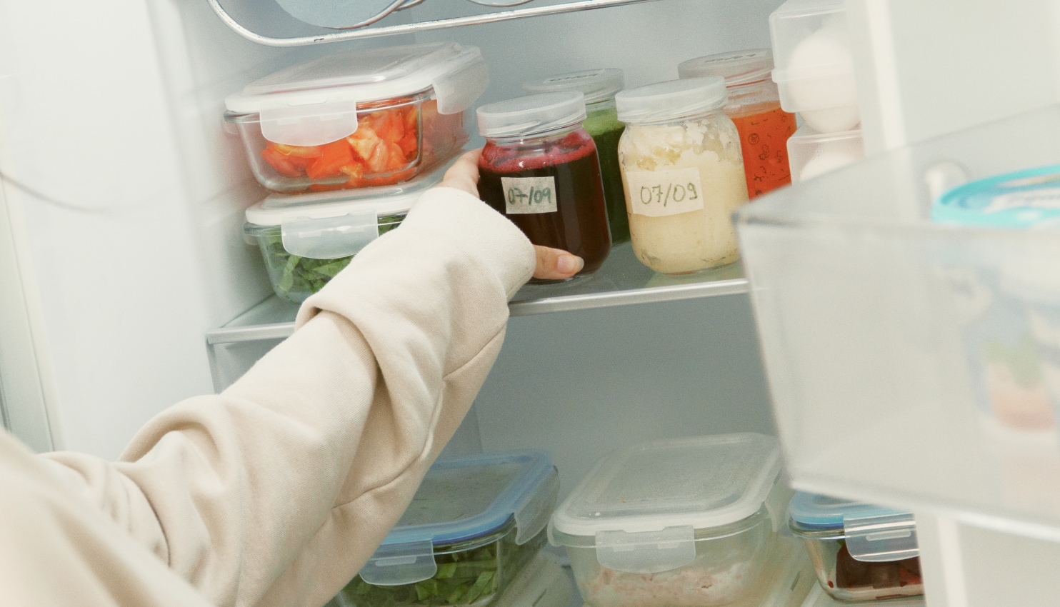 Una madre sostiene un frasco de vidrio identificado con una fecha, dentro de un refrigerador organizado con recipientes de alimentos.