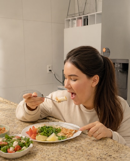 Una mamá prueba la comida con una cuchara en la cocina mientras lleva a su bebé en un portabebés.