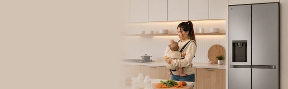 Una madre sostiene a su bebé mientras prepara alimentos en una cocina moderna, con una refrigeradora LG de puertas francesas al fondo.
