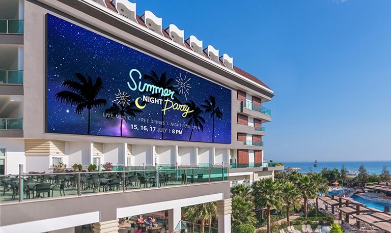 Se observa un LED grande en la pared exterior del edificio junto a la piscina al aire libre de un resort junto a la playa, y muestra claramente el anuncio del evento del resort.