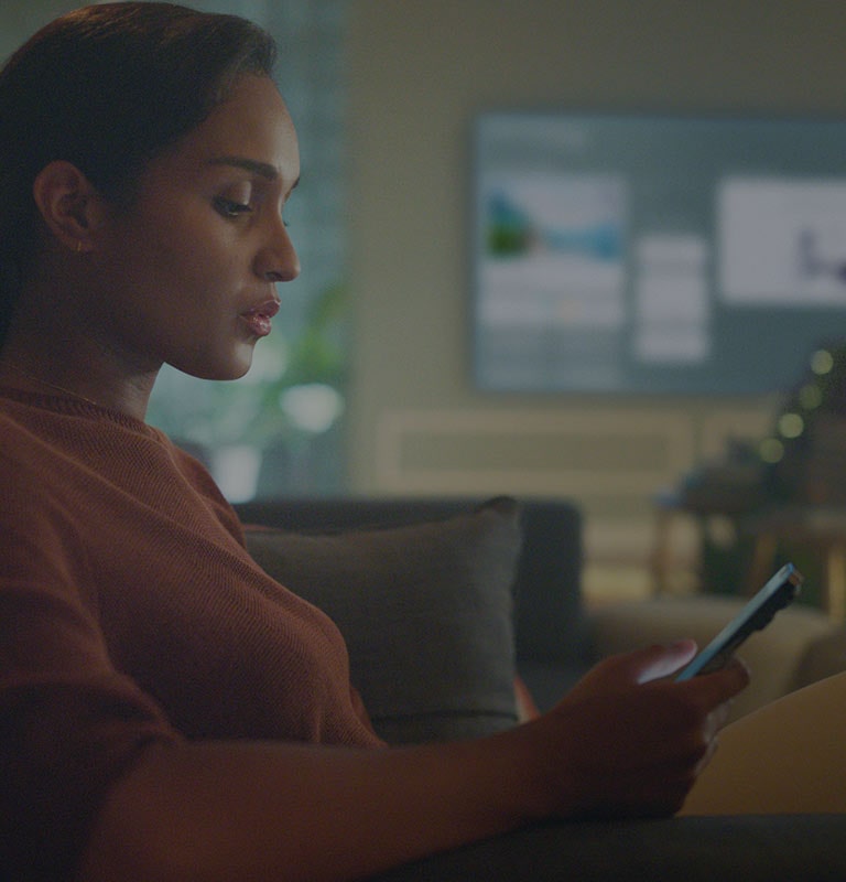 La imagen muestra a una mujer mirando su teléfono en el sofá de su sala mientras la televisión muestra el Homeboard de fondo.