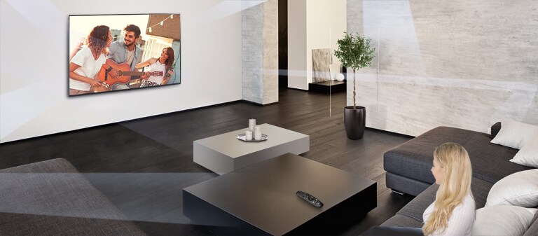 Una vista amplia de una habitación luminosa, con un televisor montado en una pared y una mujer sentada en un sofá contra la pared opuesta mirando el televisor.