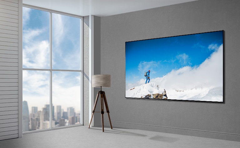 Una imagen de un televisor de pantalla plana montado en una pared gris junto a una ventana que va del piso al techo y muestra un paisaje urbano. Hay una lámpara a la izquierda del televisor y la pantalla muestra a un excursionista sobre una roca cubierta de nieve contra un cielo azul con nubes.