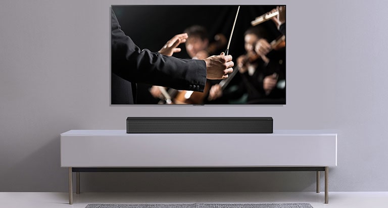 TV en una pared gris y barra de sonido LG abajo en un estante gris. La TV tiene un director dirigiendo una orquesta.