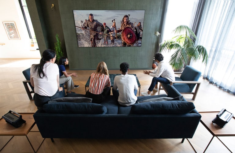 A group of friends watching a war movie on TV in the living room with 2 same Bluetooth speakers at rear speaker position