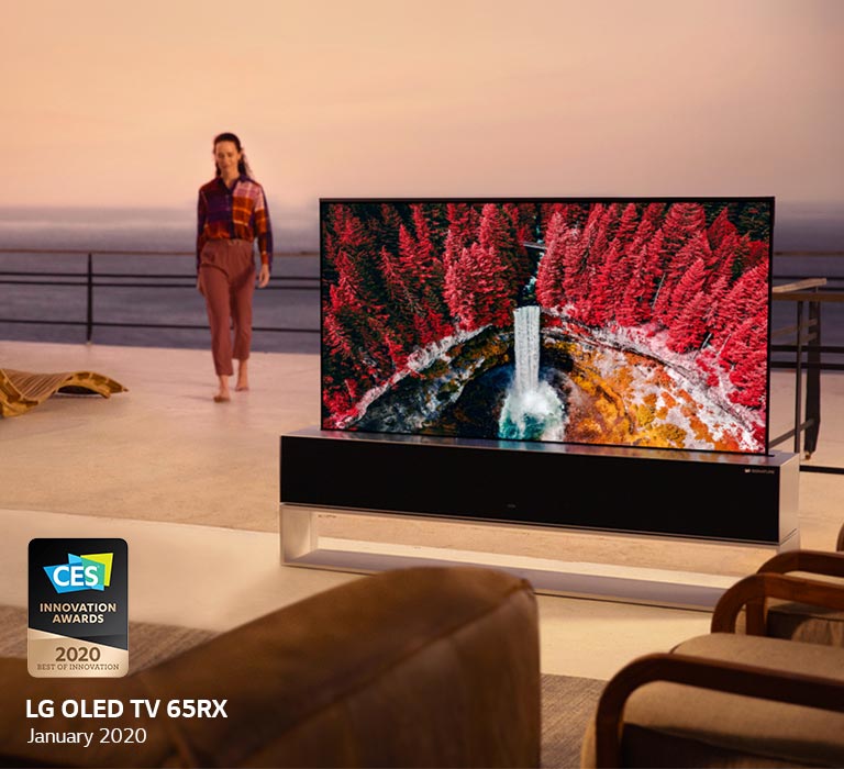A living room with a Rollable TV displaying a waterfall between maple trees, while a woman is walking inside from the terrace
