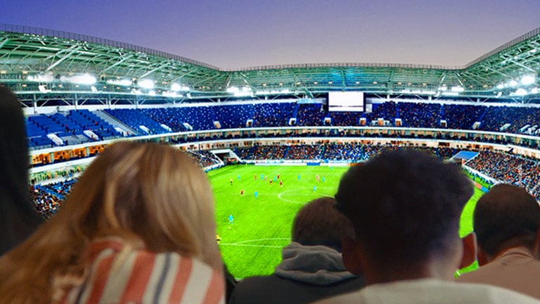 People are watching a soccer game, with a backdrop of the entire stadium, on a large TV