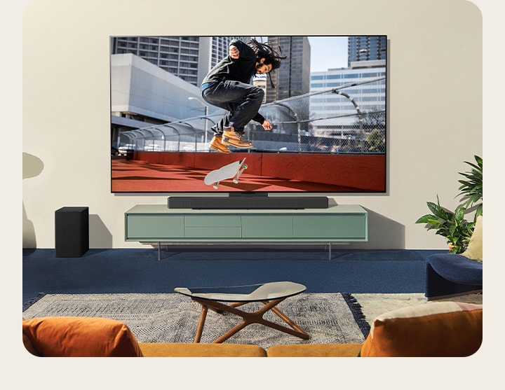 Two women sitting on a sofa in front of a wall-mounted LG TV in a colorful living room, and one woman points a remote at the TV screen. 	