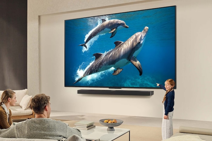 A family sits on a couch facing a LG QNED TV mounted on a wall above a LG Soundbar with a little girl pointing to a screen depicting two dolphins. The title talks about how every action is more thrilling on a massive screen.