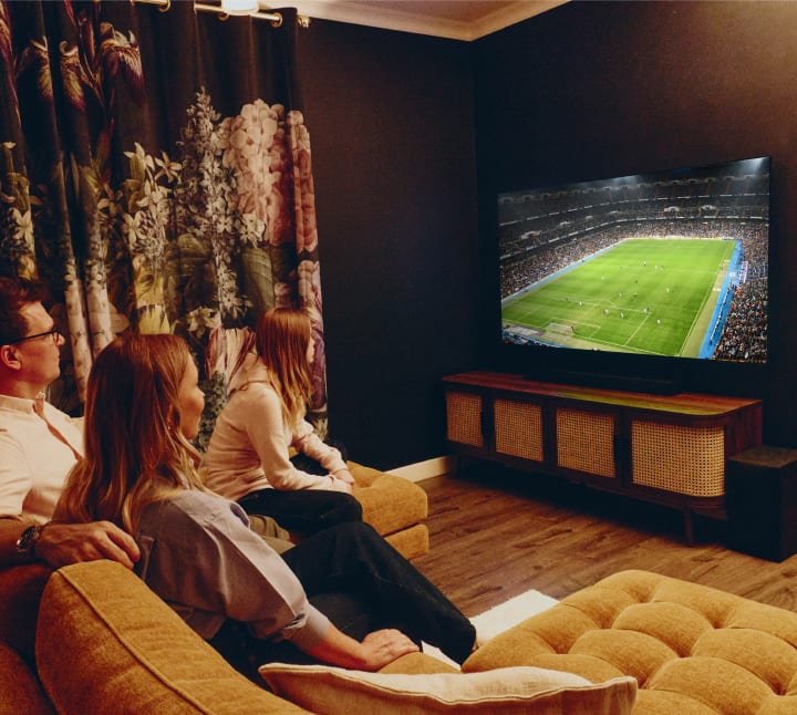 Trzysób rodzina siedzi razem na kanapie w przytulnym, przyciemnionym salonie, oglądając mecz piłki nożnej na dużym telewizorze LG OLED. Pokój ma ciemne ściany, floralne zasłony, drewnianą konsolę medialną i głośniki, tworząc ciepłą i intymną atmosferę.