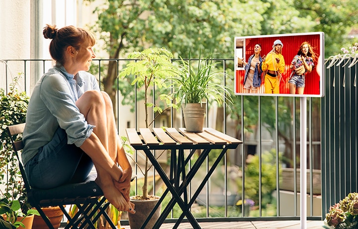 Kobieta siedzi na zewnętrznym balkonie i ogląda treści na telewizorze LG StanbyME 2 umieszczonym na regulowanym, rolowanym stojaku.