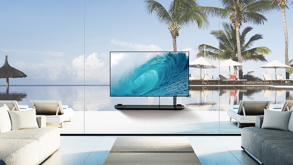 LG SIGNATURE OLED TV W pokazuje chłodną falę na ekranie, gdy jest układany w salonie z widokiem na niebieski ocean za oknem.