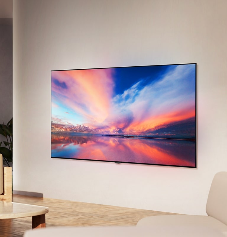 LG OLED TV, OLED B4 na ścianie neutralnego salonu, wyświetlający kolorowe zdjęcie zachodu słońca nad oceanem.