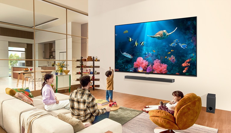 Rodzina będąca w salonie z bardzo dużym telewizorem LG, zamontowanym na ścianie, ze sceną oceanu z koralowcami i żółwiem na ekranie.