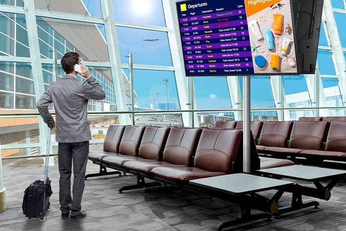 Un voyageur se tient devant un tableau d’informations de vol de l’aéroport par une journée ensoleillée, regardant les informations affichées.