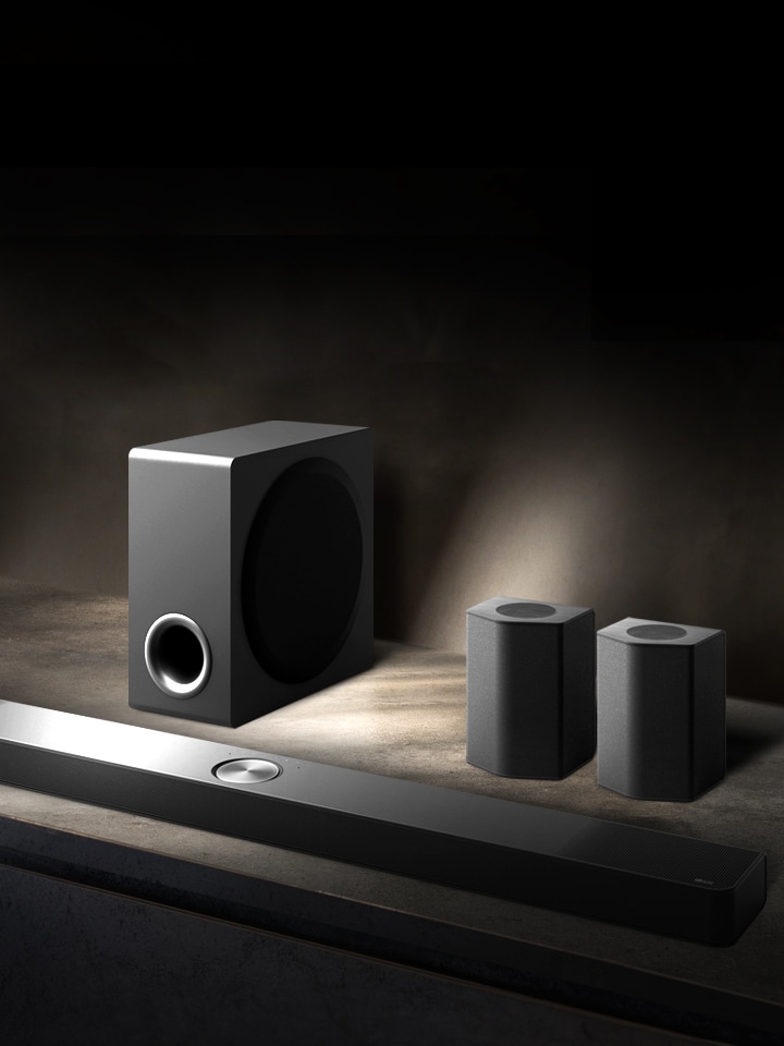 A LG Soundbar, as colunas traseiras e o Subwoofer estão colocados numa perspetiva em ângulo sobre uma prateleira de madeira castanha numa sala preta, envolta em escuridão com a luz a incidir apenas sobre o sistema de som.