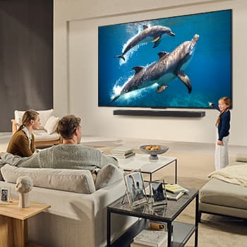Uma família sentada num sofá virada para uma LG QNED TV montada na parede por cima de uma LG Soundbar com uma menina a apontar para um ecrã onde vemos dois golfinhos. O título fala de como cada ação é mais emocionante num ecrã gigante.