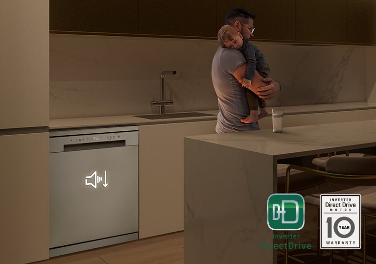 Homem a segurar num bébé numa cozinha pouco iluminada, com a máquina de loiça a funcionar silenciosamente no fundo.