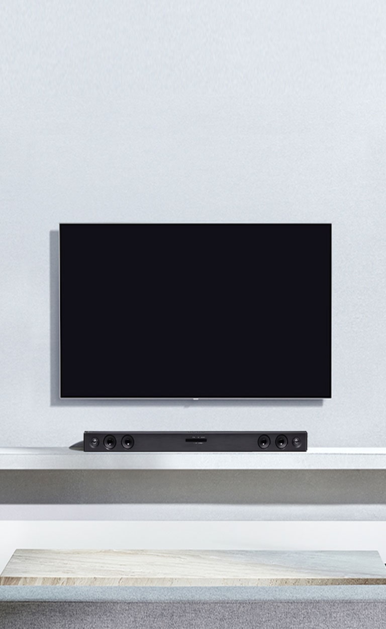 A TV LG está pendurada na parede cinzenta. E a barra de som LG SQC2 está colocada na prateleira na sala de estar. E o subwoofer está colocado ao lado da prateleira. A imagem ilustra que a barra de som corresponde à TV LG e complementa o interior.
